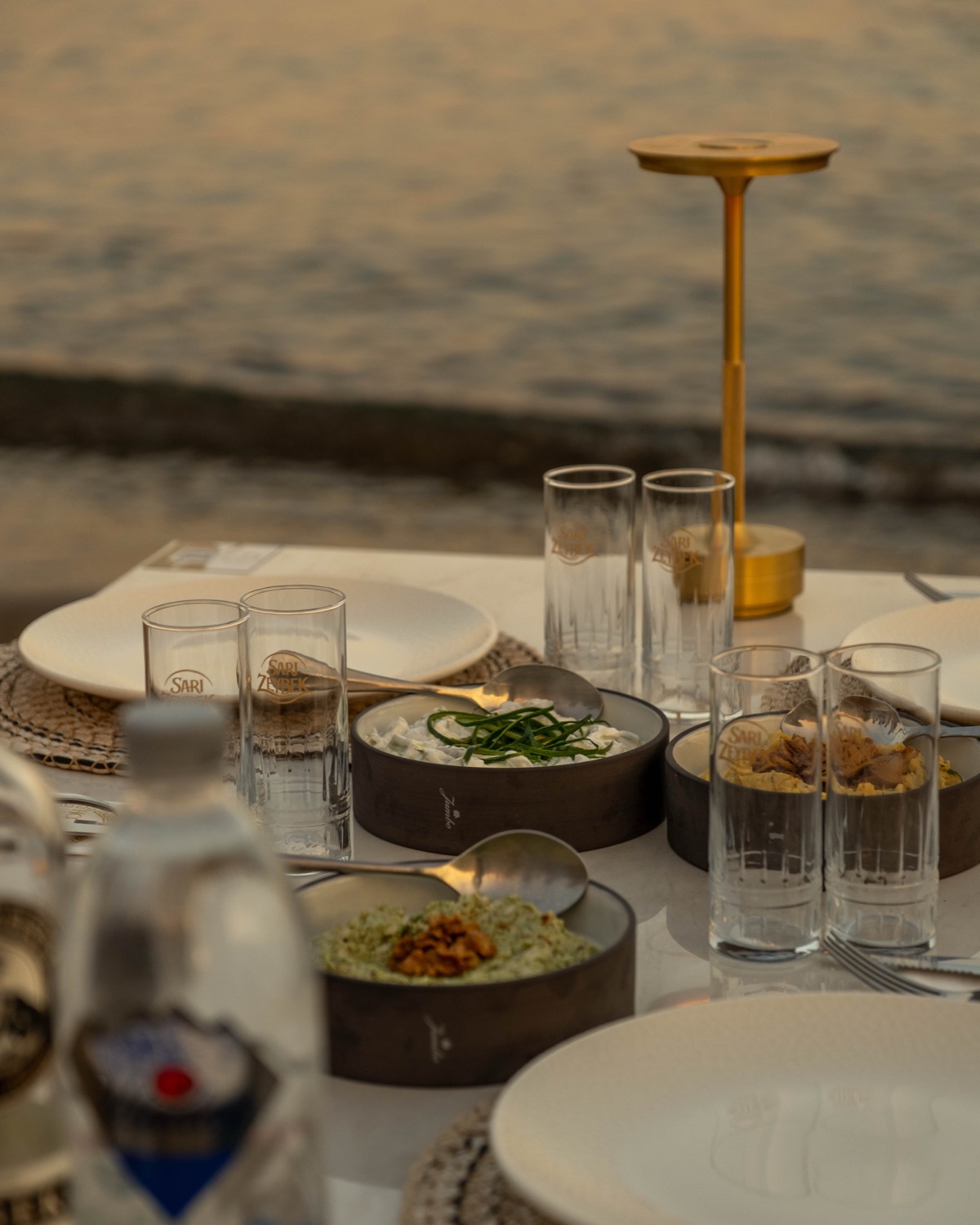 Altın masa lambası, kristal bardaklar ve mezeler; arkada akşam ışığında deniz